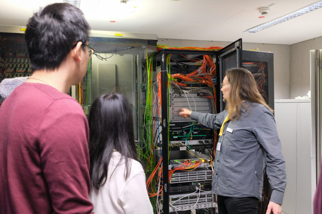 Ein Mann steht vor einem geöffneten Server-Schrank. Er deutet auf die darin verlaufenden Kabelstränge. Links im Bild stehen zwei weitere Personen und schauen mit etwas Abstand in den geöffneten Server-Raum.