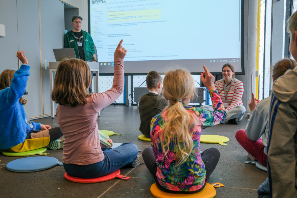 Im Vordergrund sitzt eine Gruppe jüngerer Kinder auf bunten Sitzkissen auf dem Boden. Sie melden sich. Im Hintergrund sitzt eine erwachsene Frau  ihnen gegenüber und lächelt. An der Wand hinter ihr wird ein Bild des Suchfensters des DNB-Kataloges auf eine Leinwand geworfen. Links daneben steht ein Mann an einem Computer.