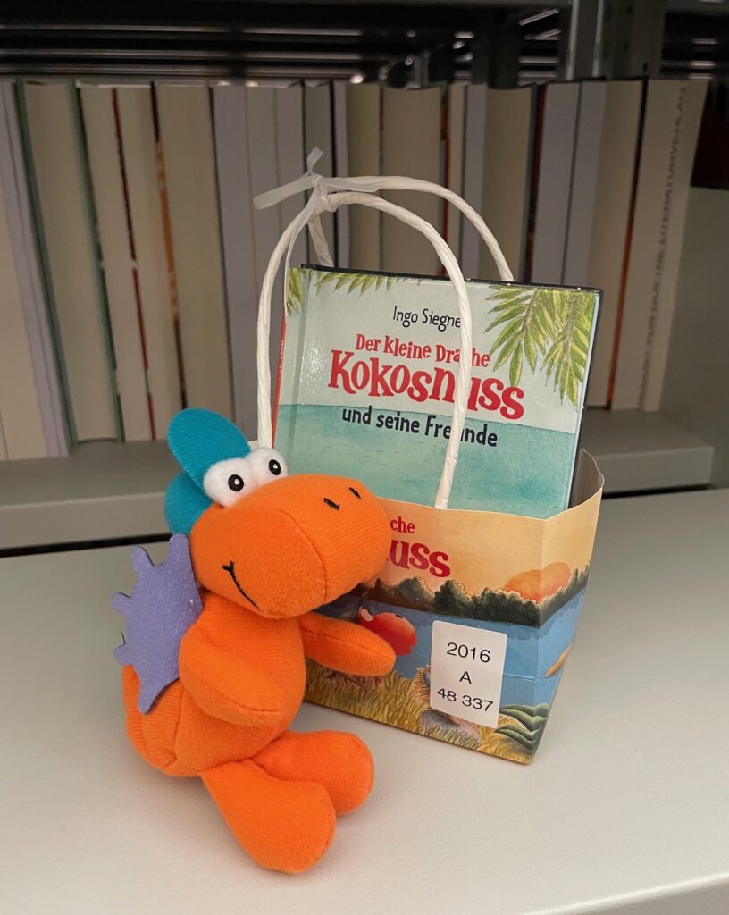 Orange-lila Plüschdrache mit Buch und einer Tasche in einem Bücherregal