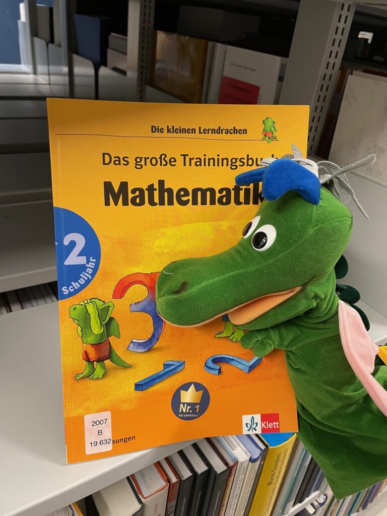 Grüne Drachen-Handpuppe mit einem Mathamatikbuch vor einem Bücherregal
