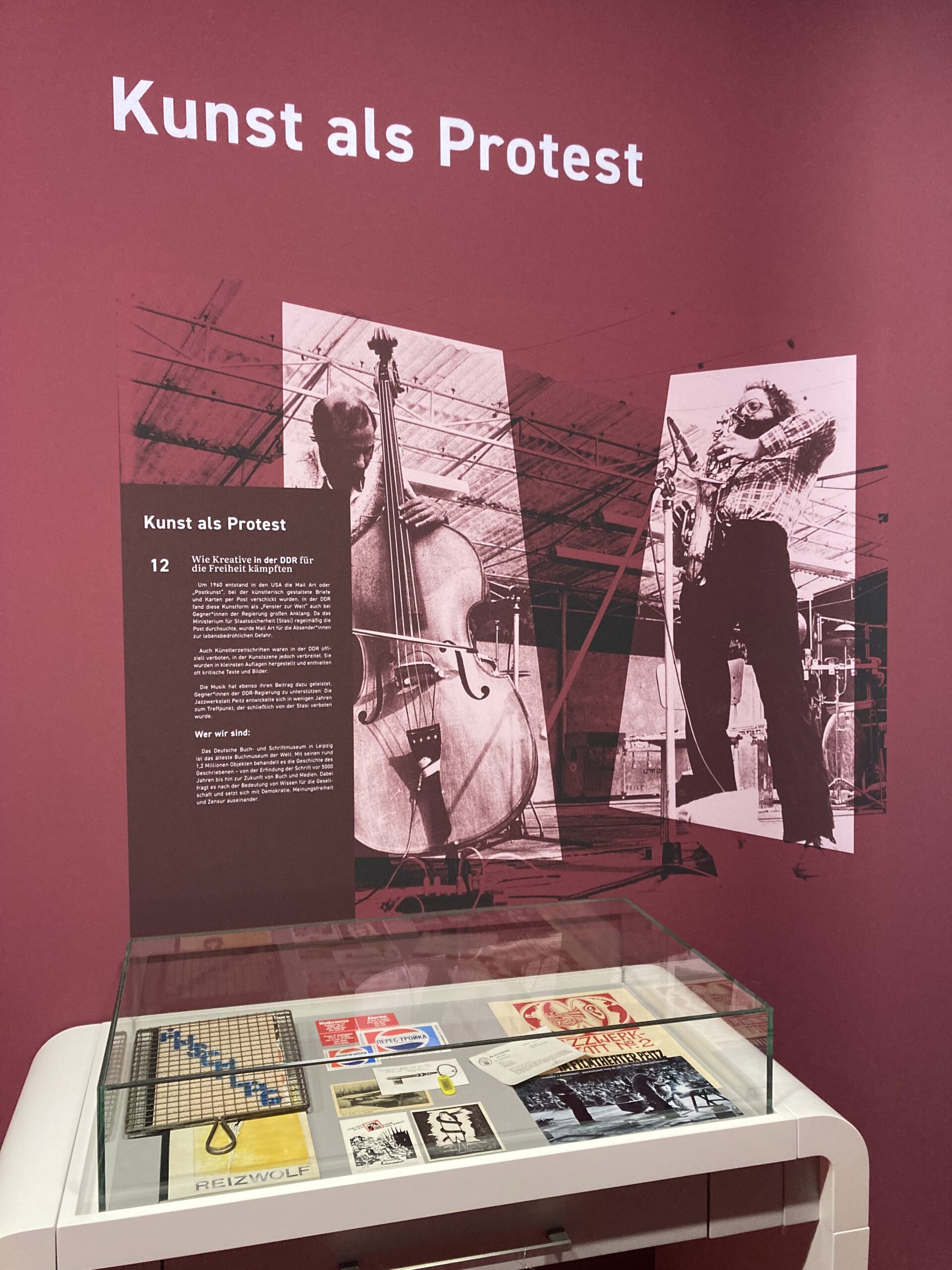 Auf einer roten Wand sind Bilder und Text aufgebracht. Davor steht eine kleine Vitrine mit ausgestellten Büchern und Postkarten.