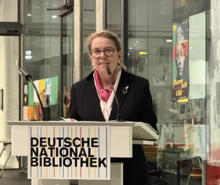 Eine Frau im Anzug, steht an einem Rednerpult und spricht ins Mikrofon.