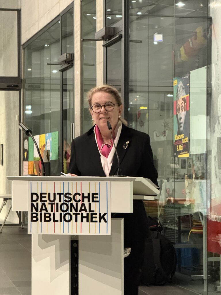 Eine Frau im Anzug, steht an einem Rednerpult und spricht ins Mikrofon.