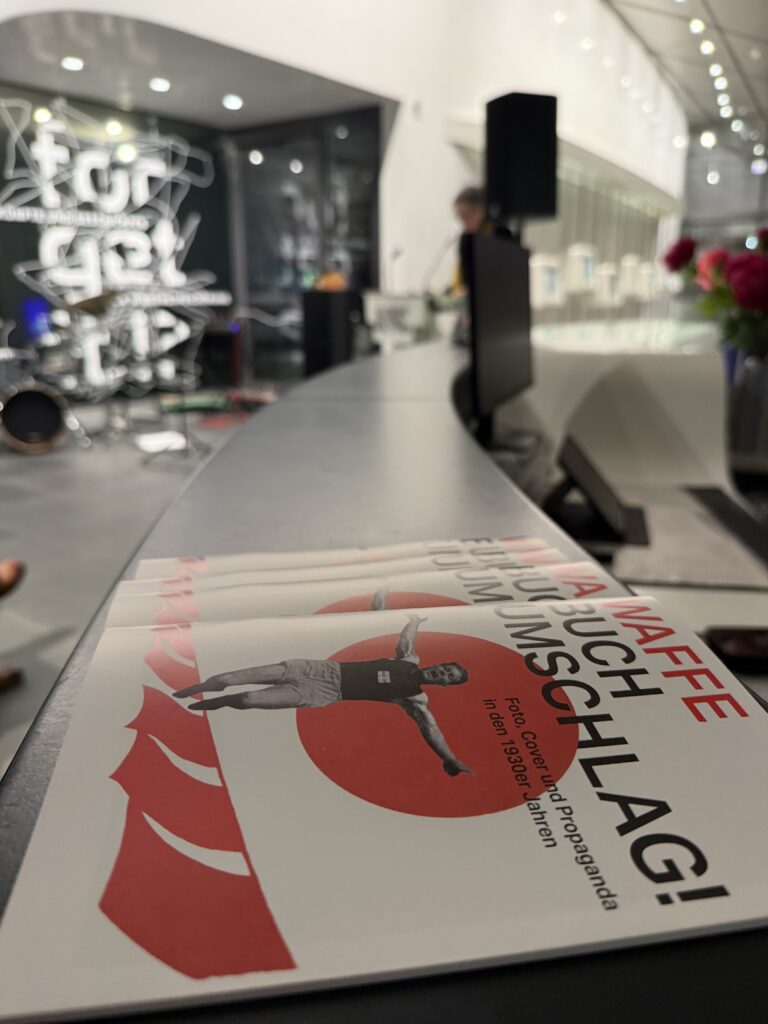 Im Vordergrund liegen mehrere Booklets der Ausstellung ‚Waffe Buch Umschlag!‘ übereinander.