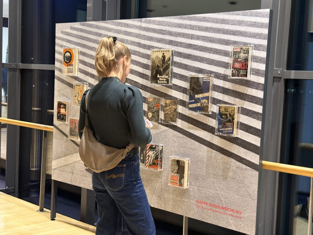 Eine Frau, die man nur von hinten sieht, blickt auf eine Ausstellungswand, an der mehrere Bücher befestigt sind.