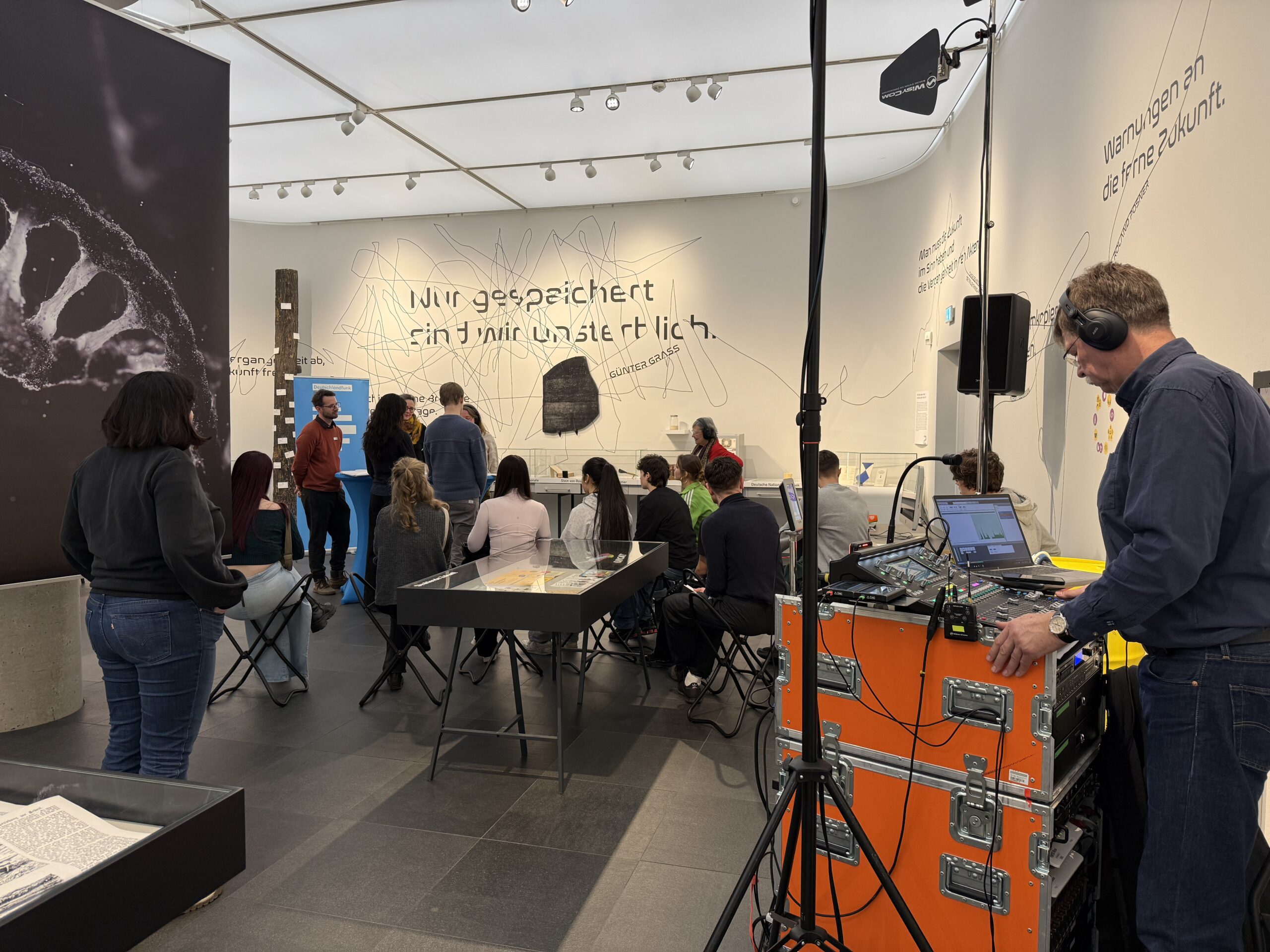 In einer Ausstellung sitzen viele Menschen in einem Gespräch, im Vordergrund sieht man einen Tontechniker an seinem Mischpult.