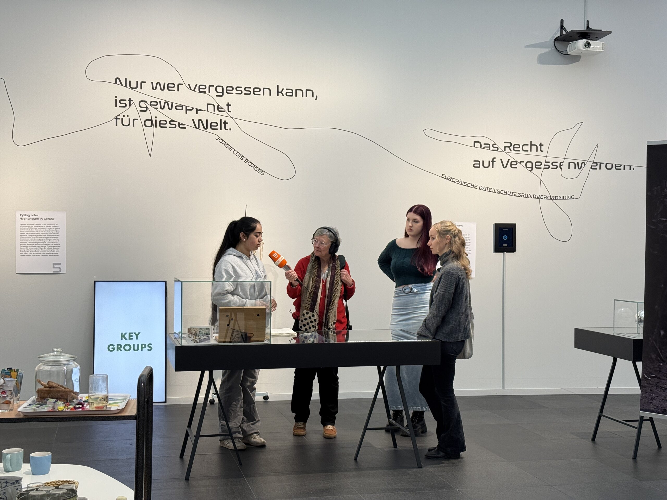 Eine Frau mit Kopfhörern und Mikrophon interviewt drei junge Menschen in einer Ausstellung.