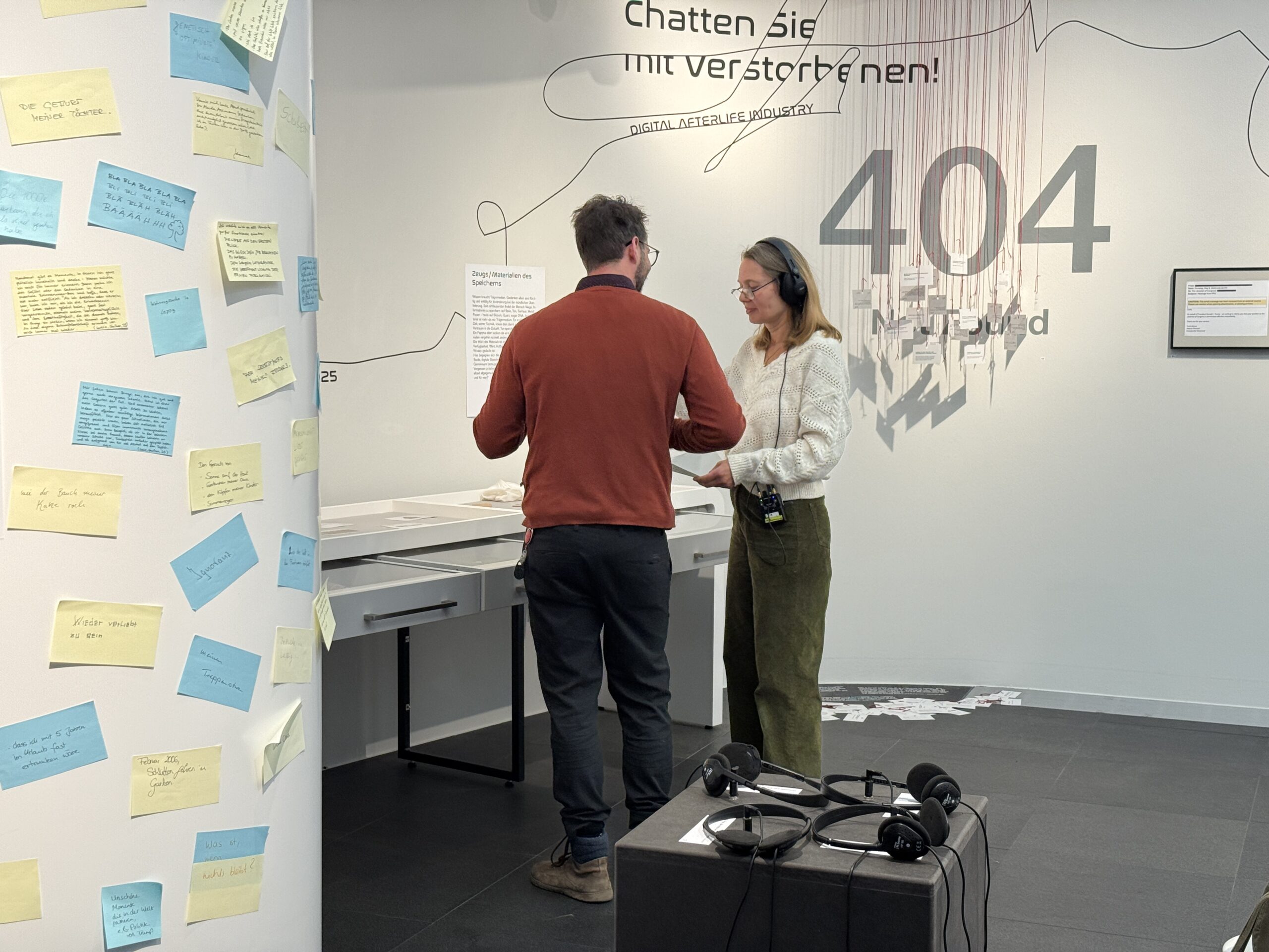 Eine Frau mit Kopfhörern und ein Mann unterhalten sich in einer Ausstellung. Im Vordergrund ist eine Säule mit gelben und blauen Klebezetteln zu sehen.