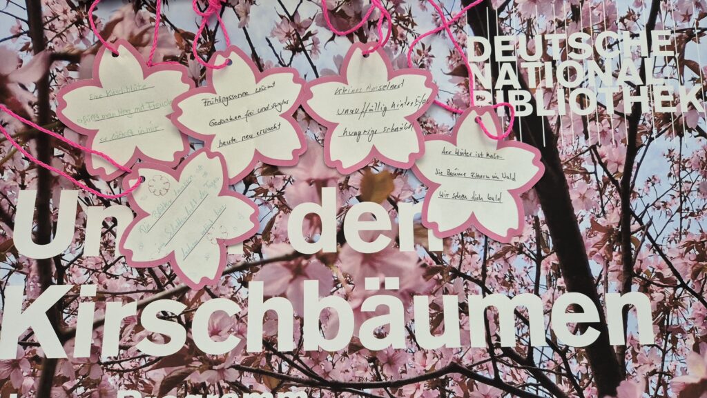 Auf einem rosa Plakat liegen Papiere in Kirschblütenform. Auf sie sind japanische Gedichte geschrieben.