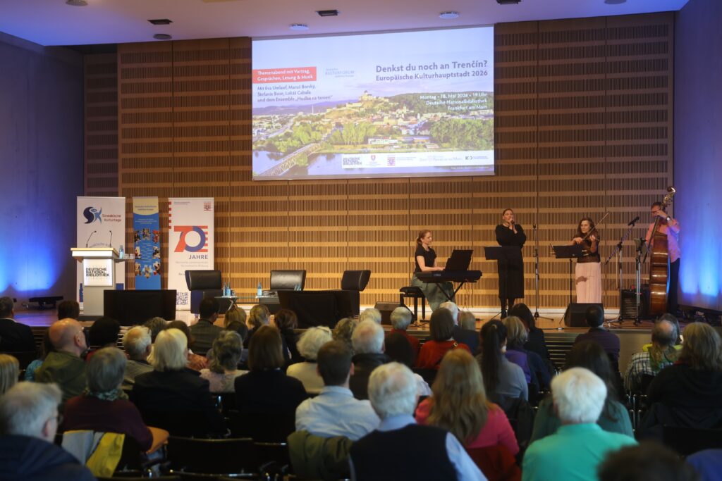 Der Veranstaltungsaal der Deutschen Nationalbibliothek in Frankfurt am Main ist gut gefüllt. Auf der Bühne steht links ein Rednerpult hinter dem verschiedene Roll-Ups, unter anderem der Solwakischen Kulturtage, stehen. Auf der Leinwand in der Mitte wird eine Präsentation gezeigt. Man sieht den Titel der Veranstaltung "Denkst du noch an Trencin" vor einem Bild der slowakischen Stadt. Darunter der Zusatz: Europäische Kulturhauptstadt 2026. In der rechten Bildhälfte steht eine Gruppe Musikerinnen an verschiedenen Instrumenten. Eine Frau ist Sängerin und hält ein Mikrofon in der Hand