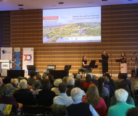 Der Veranstaltungsaal der Deutschen Nationalbibliothek in Frankfurt am Main ist gut gefüllt. Auf der Bühne steht links ein Rednerpult hinter dem verschiedene Roll-Ups, unter anderem der Solwakischen Kulturtage, stehen. Auf der Leinwand in der Mitte wird eine Präsentation gezeigt. Man sieht den Titel der Veranstaltung "Denkst du noch an Trencin" vor einem Bild der slowakischen Stadt. Darunter der Zusatz: Europäische Kulturhauptstadt 2026. In der rechten Bildhälfte steht eine Gruppe Musikerinnen an verschiedenen Instrumenten. Eine Frau ist Sängerin und hält ein Mikrofon in der Hand