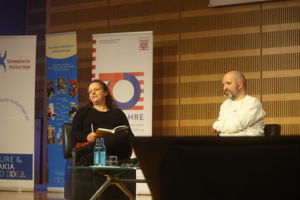 Lukás Cabala mit der Übersetzerin Stefanie Bose. Diese hat ein aufgeschlagenes Buch auf dem Schoss. Beide sitzen auf einer Bühne und schauen ins Publikum.