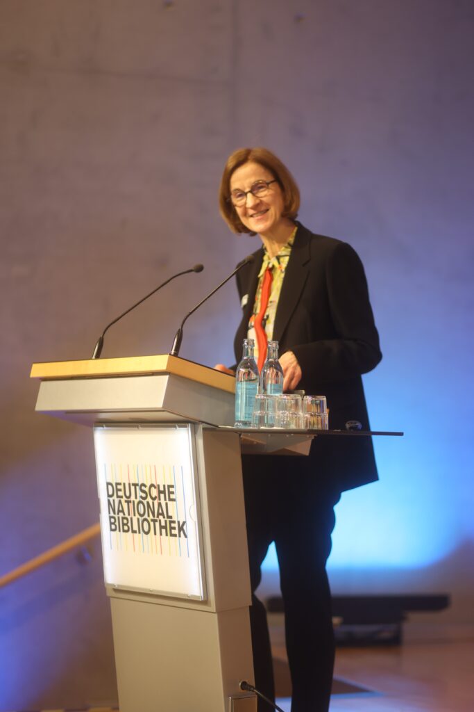 Sylvia Asmus, die Direktorin des Deutschen Exilarchivs der Deutschen Nationalbibliothek in Frankfurt am Main steht an einem Rednerpult und blickt den Betrachter mit einem sympathischen Lächeln entgegen.