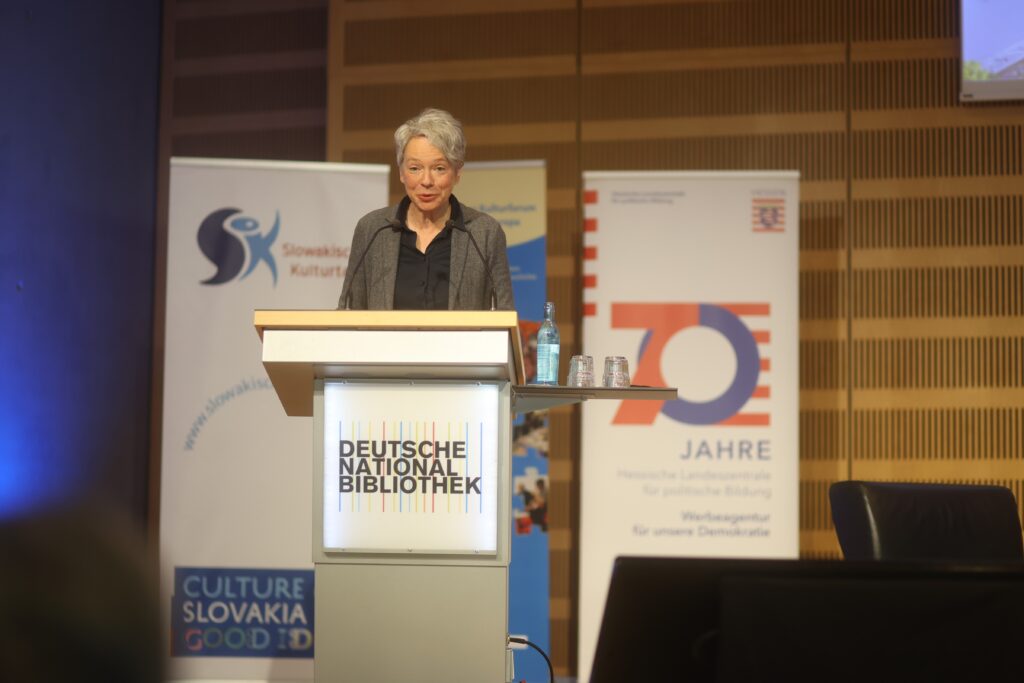 Dr. Ina Hartwig die Kulturdozentin der Stadt Frankfurt am Main steht an einem Rednerpult mit dem Logo der Deutschen Nationalbibliothek. Sie sieht den Betrachter mit einem zugewandten, offenen Gesichtsausdruck an.