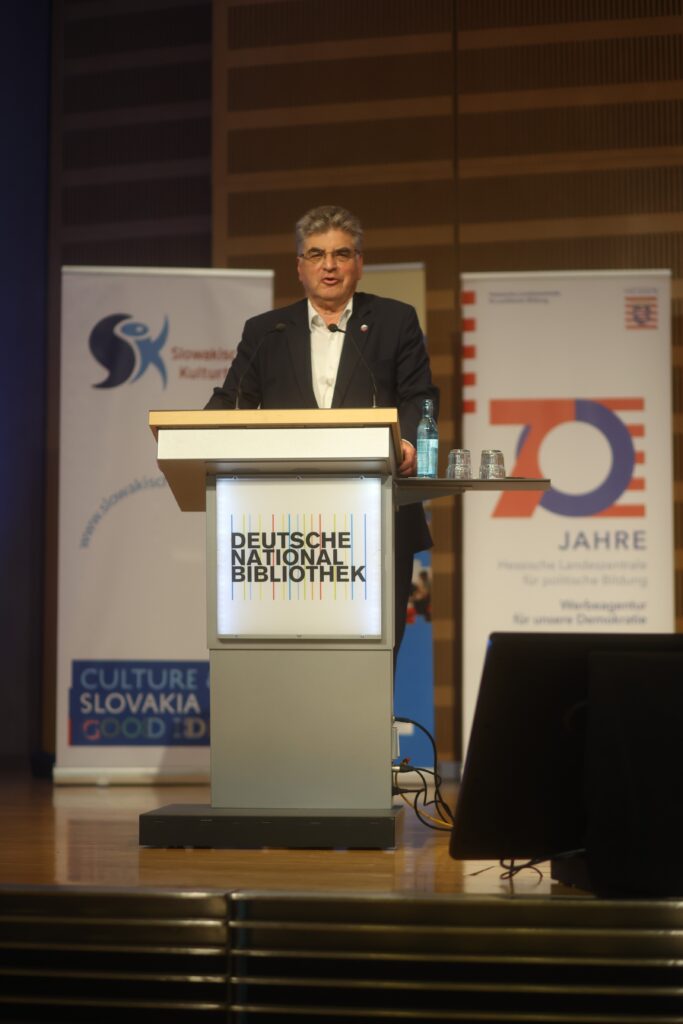 Imrich Donath, der Honorarkonsul der Slowakischen Republick steht an einem Rednerpult mit dem Logo der Deutschen Nationalbibliothek. Er sieht den Betrachter direkt an.