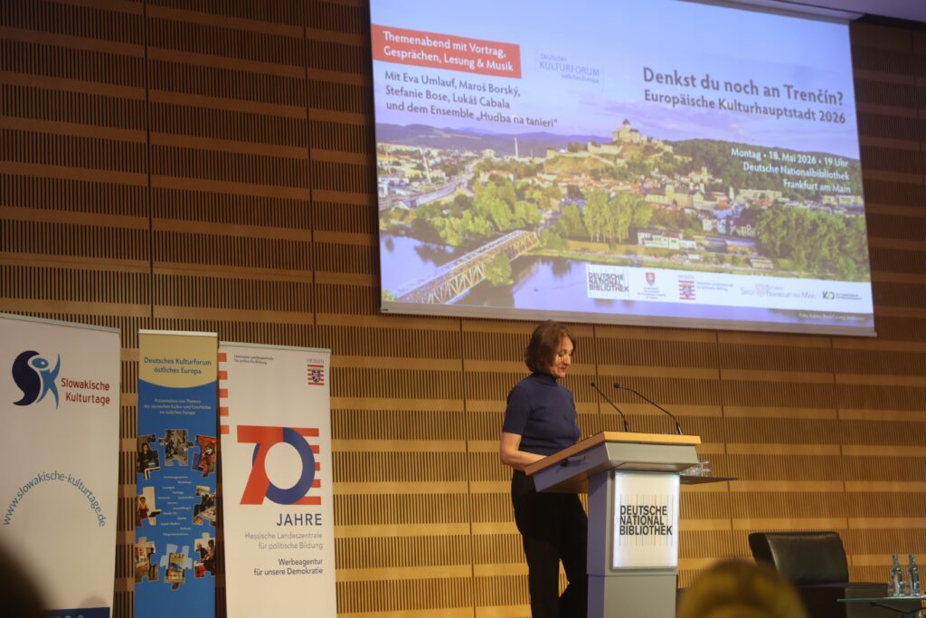 Tanja Krombach die stellvertretenden Direktorin des Deutschen Kulturvorums östliches Europa steht an einem Rednerpult mit dem Logo der Deutschen Nationalbibliothek. Im Hintergrund eine Leindwand mit einer Präsentation. Sie trägt den Titel: "Denkst du noch an Trencin?" Hinter dieser Überschrift sieht man ein Foto der Stadt Trencin.