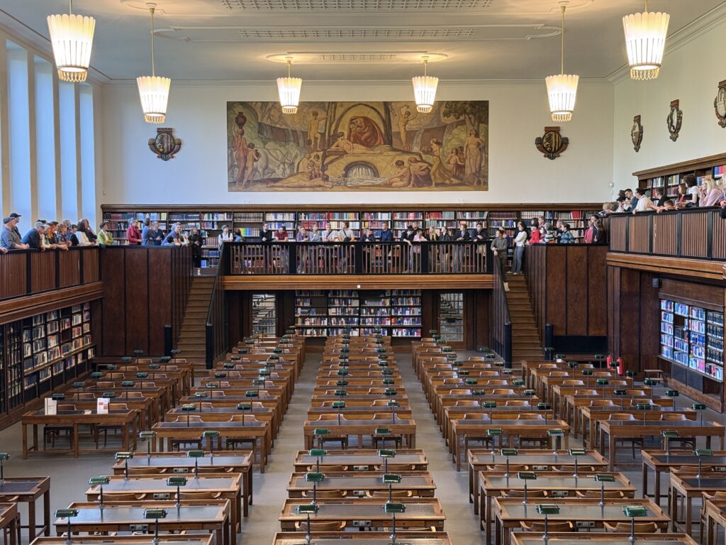 Blick in einen großen Lesesaal. An der Decke hängen historische Lampen. Auf der Gallerie stehen viele Menschen.