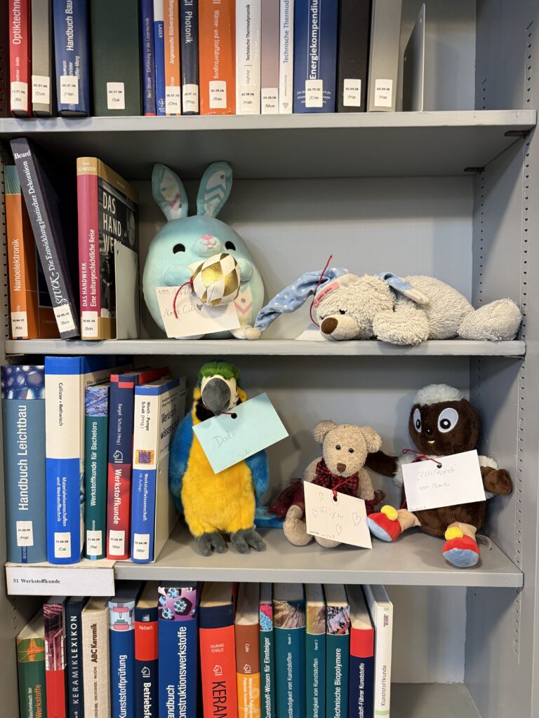 Mitten in einem Regal mit Büchern sitzen fünf Kuscheltiere, darunter zwei Bären, Pittiplatsch, ein Papagei und ein türkiser Hase.