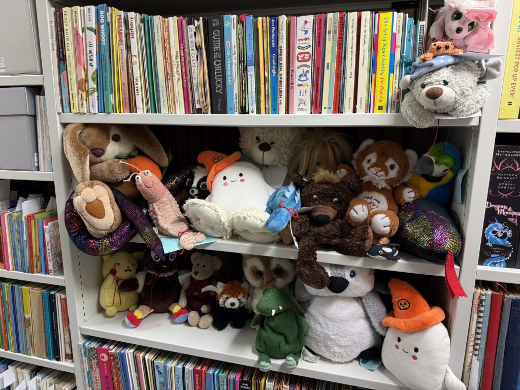 In einem Regal mit bunten Buchrücken sitzen unzählige Kuscheltiere