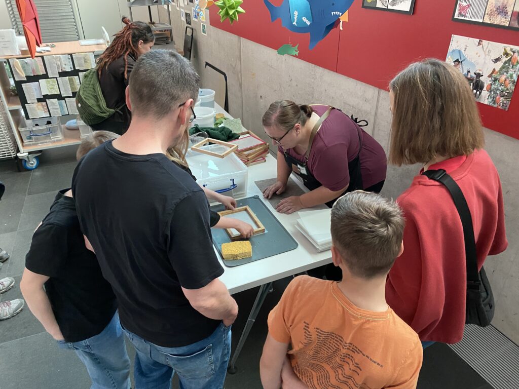 Menschen stehen um einen Tisch. Eine Frau zeigt, wie Papier in einen Rahmen geschöpft wird.