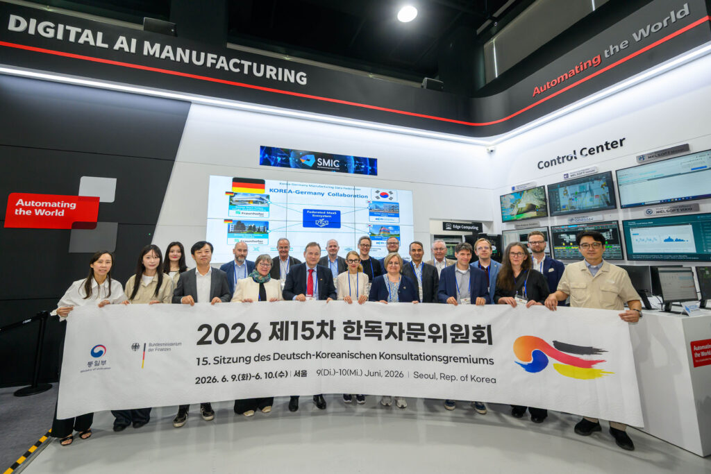 Mitarbeitende der Firma Smart Manufacturing und Teilnehmende der Konferenz halten ein Banner der 15. Sitzung des Deutsch-Koreanischen Konultationsgremiums