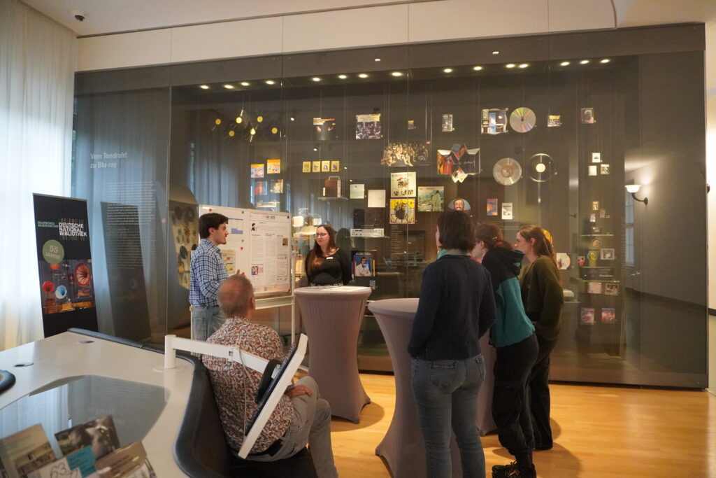 Mehrere Personen stehen in eine Ausstellung zu Tonträgern. Zwei Personen stehen vor einer Tafel und unterhalten sich. Der Rest steht an Stehtischen und hört ihnen zu