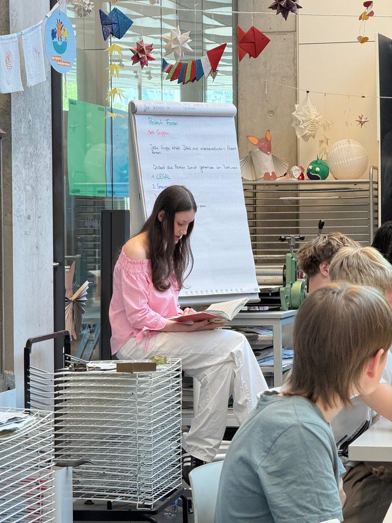 Charlotte liest aus einem Buch, vor ihr sitzen mehrere Jugendliche. 