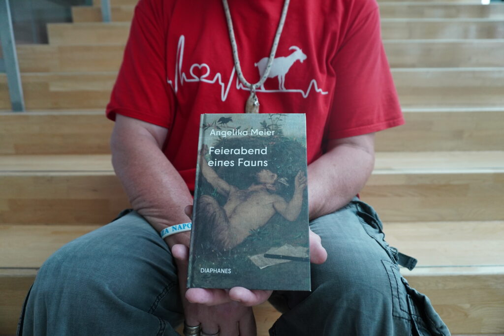 Auf einer Treppe sitzt ein Mann und hält ein Buch in die Kamera. Der Bildausschnitt ist so gewählt, dass man nur seinen Oberkörper und seine Beine bis zu den Knien sieht.