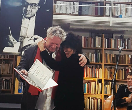Ein Mann und eine Frau umarmen sich. Erhält ein Buch in der Hand, im Hintergrund ist ein Bücherregal.