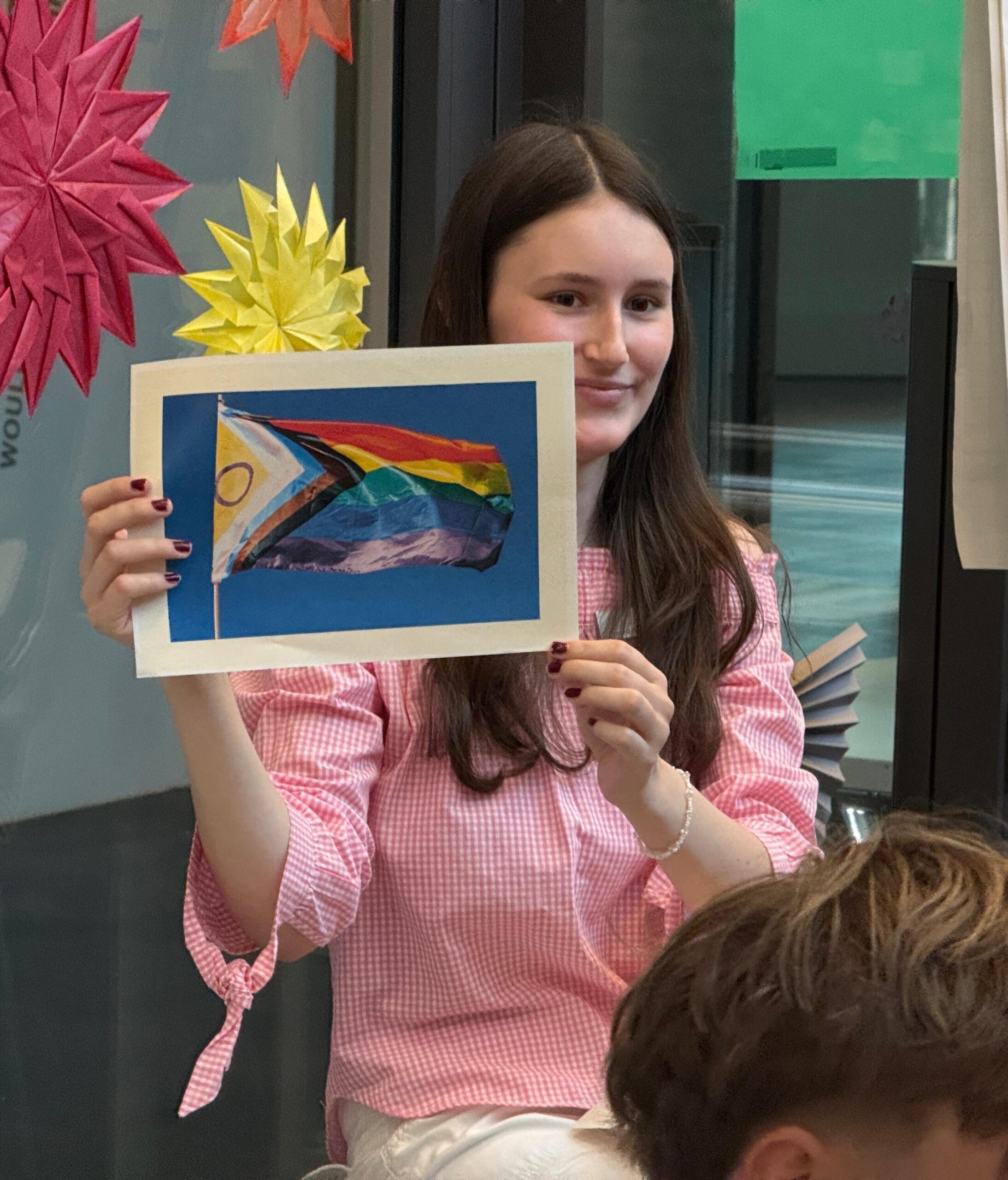 Charlotte zeigt ein Bild mit einer LGBTQ+ Flagge hoch.