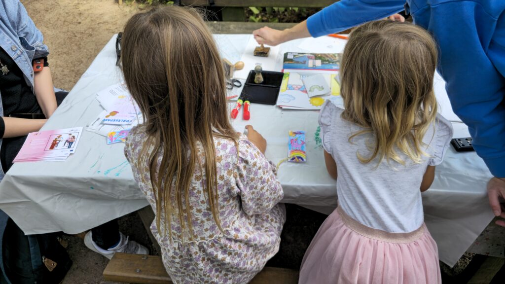 Auf einem Tisch liegen Materialien zum Basteln, unter anderem Klebestifte, Stempel und Ausmalbilder. Davor sitzen zwei Kinder und basteln.