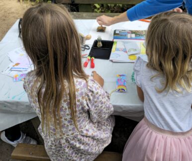 Auf einem Tisch liegen Materialien zum Basteln, unter anderem Klebestifte, Stempel und Ausmalbilder. Davor sitzen zwei Kinder und basteln.