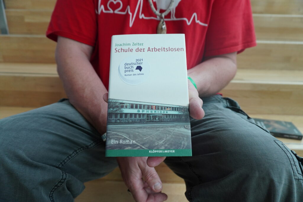 Auf einer Treppe sitzt ein Mann und hält ein Buch in die Kamera. Der Bildausschnitt ist so gewählt, dass man nur seinen Oberkörper und seine Beine bis zu den Knien sieht.