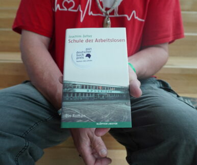 Auf einer Treppe sitzt ein Mann und hält ein Buch in die Kamera. Der Bildausschnitt ist so gewählt, dass man nur seinen Oberkörper und seine Beine bis zu den Knien sieht.