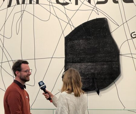 Eine Frau interviewt einen Mann, im Hintergrund ist eine große schwarze Platte an der Wand montiert (Rosetta-Stein)