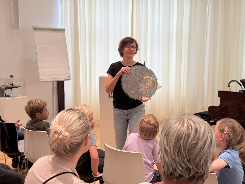 Eine Frau hält eine große Lochplatte in der Hand. Vor ihr sitzen Kinder und Erwachsene.
