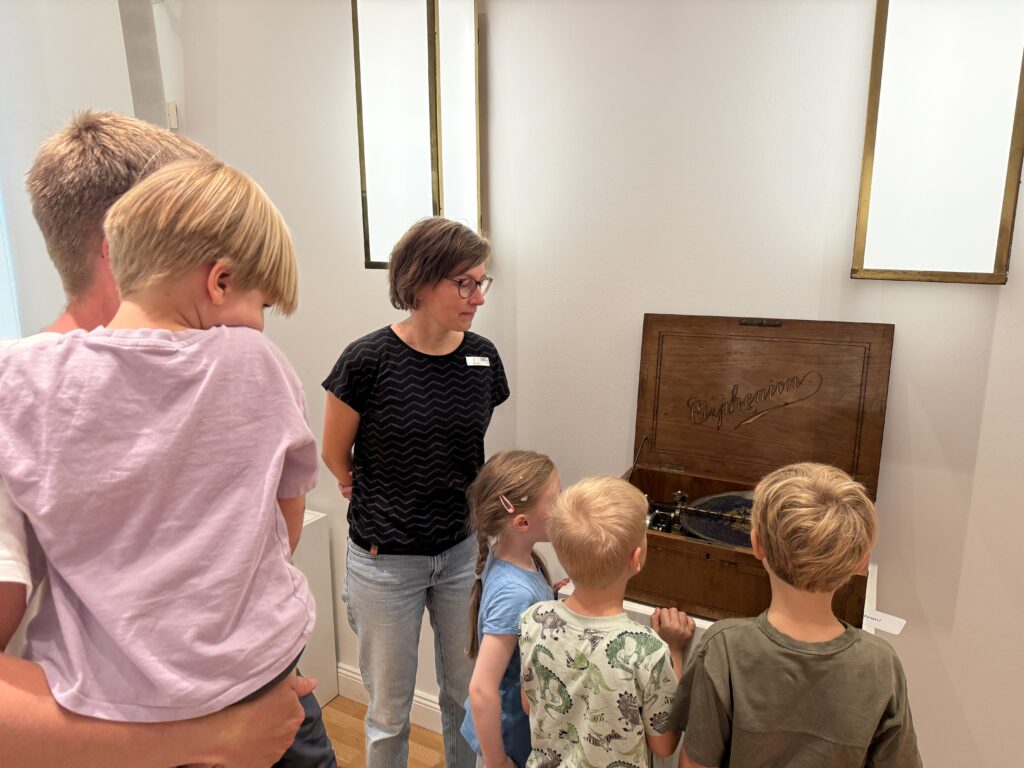 Kinder stehen um ein historisches Musikgerät und bebachten, wie sich die Lochplatte in dem Gerät dreht.