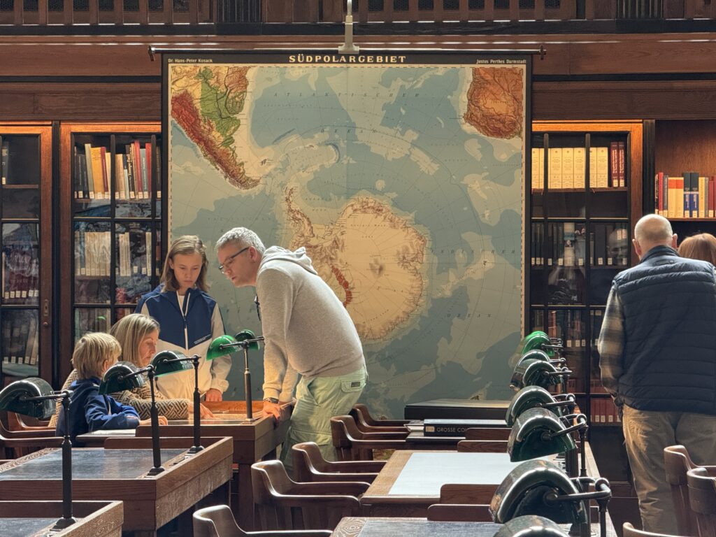 In einem historischen Lesesaal steht eine große geografische Karte. Davor sitzt eine Familie an einem Tisch und schaut sich ein Buch an.