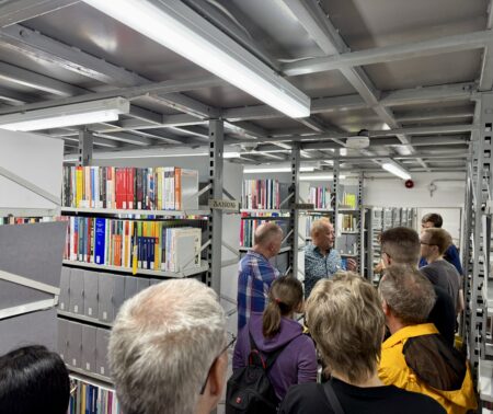 Führung in einem Magazin. Auf grauen Regalen stehen farbige Bücher. Mehrere Menschen hören zu, während ein Mann etwas erklärt.