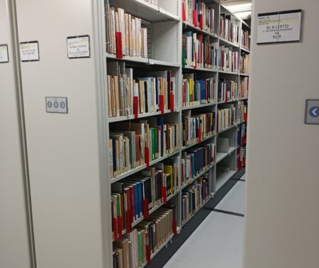 Zahlreiche Bücher füllen eine lange Regalreihe eines beweglichen Magazinschrankes. Man erkennt die hydraulischen Arme am oberen Bildrand.