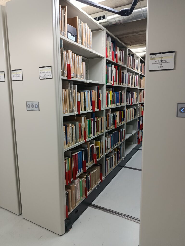 Zahlreiche Bücher füllen eine lange Regalreihe eines beweglichen Magazinschrankes. Man erkennt die hydraulischen Arme am oberen Bildrand.